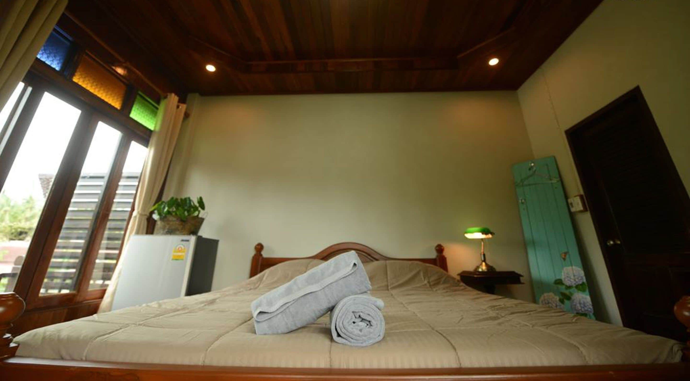 A neatly made bed with a rolled towel, a potted plant, and a lamp on a bedside table in a room with wooden ceiling beams and a door.