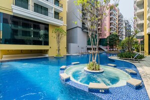 Une piscine avec un jacuzzi, entourée d’une terrasse carrelée, jouxtant un bâtiment doté de balcons et d’un escalier.