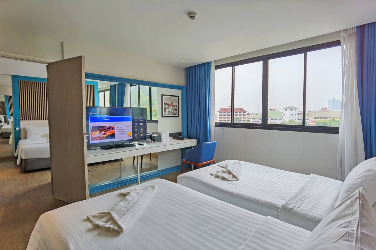 Een hotelkamer met twee bedden, een bureau met een computer, een stoel en een groot raam met gordijnen.