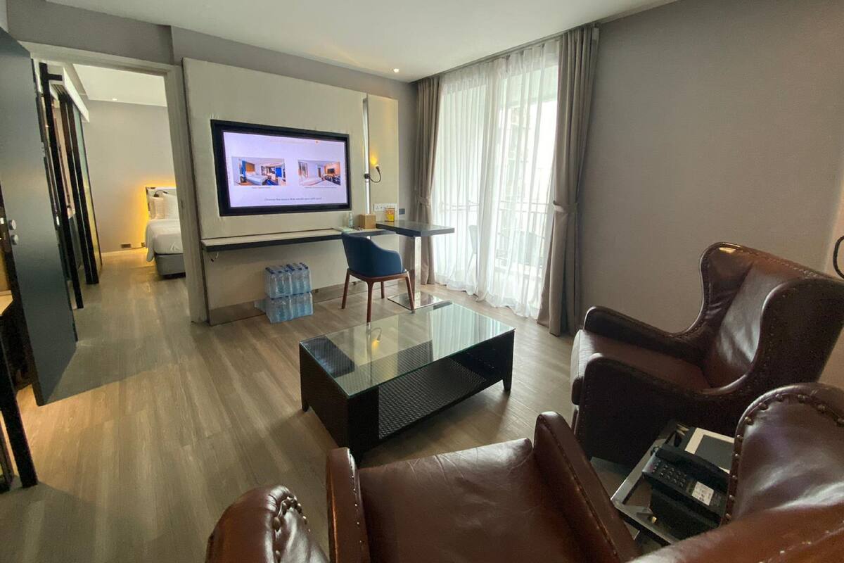 Een moderne hotelkamer met een glazen salontafel, leren fauteuils, een aan de muur bevestigde televisie en een groot raam met gordijnen.