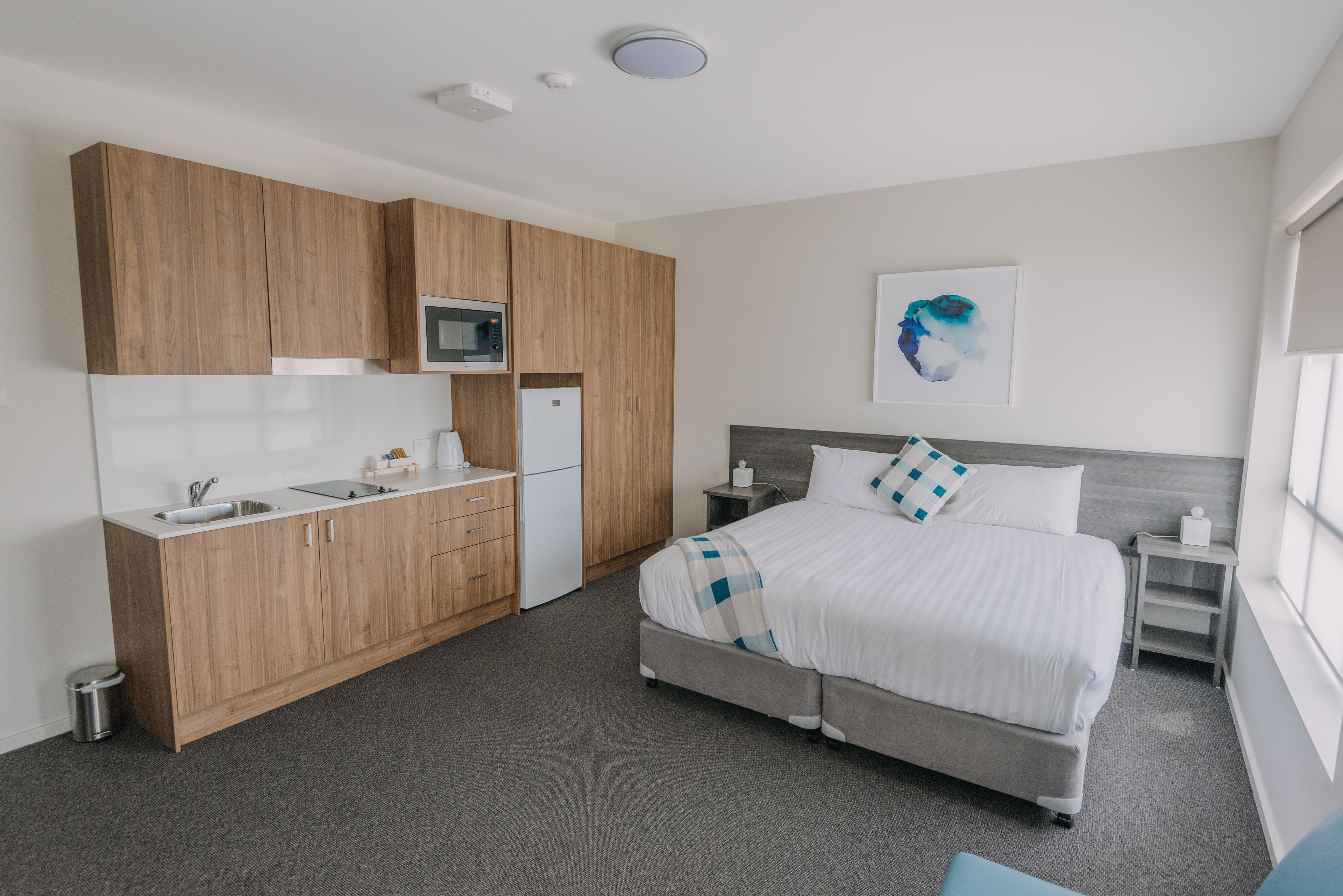 A modern hotel room with a kitchenette, a bed with a checkered blanket, a bedside table, and a wall-mounted artwork.