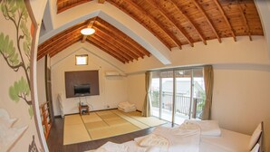 A room with a bed, a TV, a sliding glass door to a balcony, tatami mats on the floor, and wooden beams on the ceiling. - Kitakara Yuimaru in Motobu (Motobu)