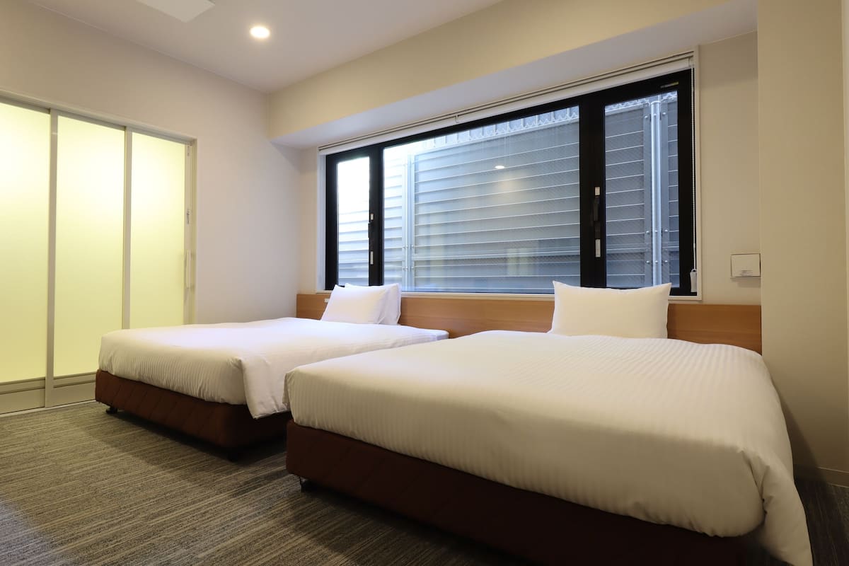 Une chambre d’hôtel avec deux lits, un bureau et une chaise. Elle est équipée d’une porte coulissante et d’une fenêtre avec des stores.