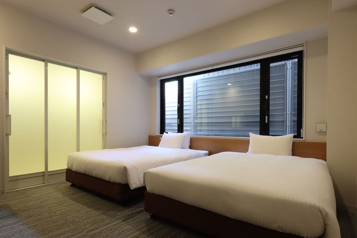 Une chambre d’hôtel avec deux lits, une porte coulissante, une fenêtre avec des stores et un sol recouvert de moquette.