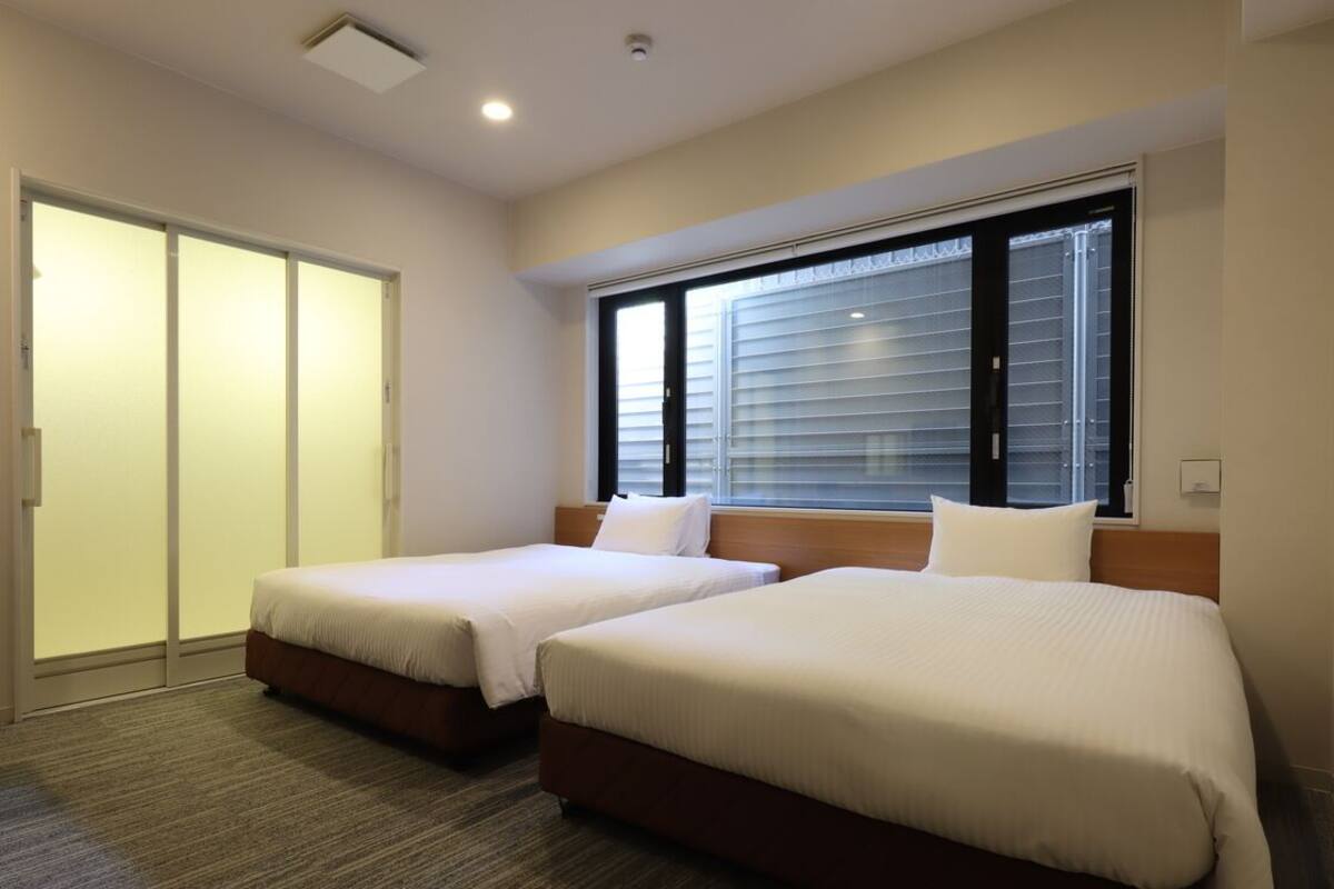Une chambre d’hôtel avec deux lits, une porte coulissante et une grande fenêtre avec des stores.