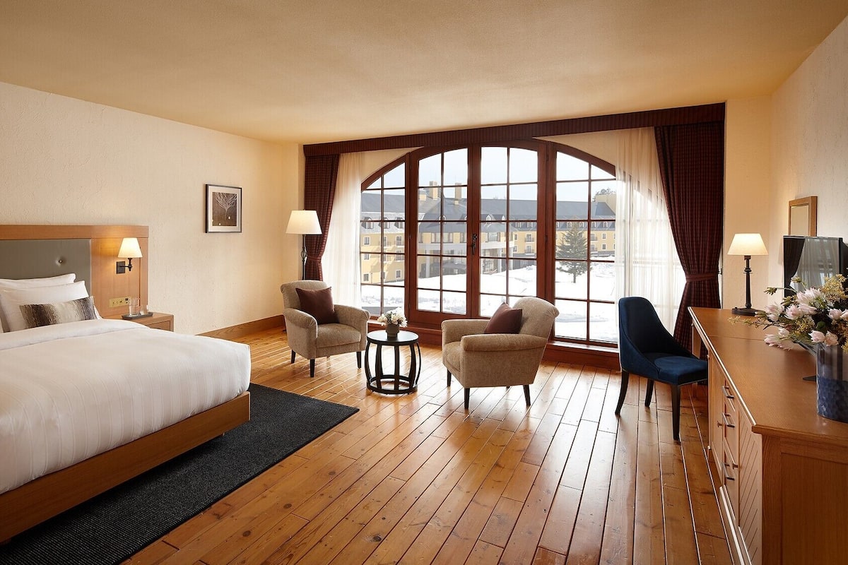 A hotel room with a large bed, two armchairs, a small table, and a desk with a lamp and flowers.