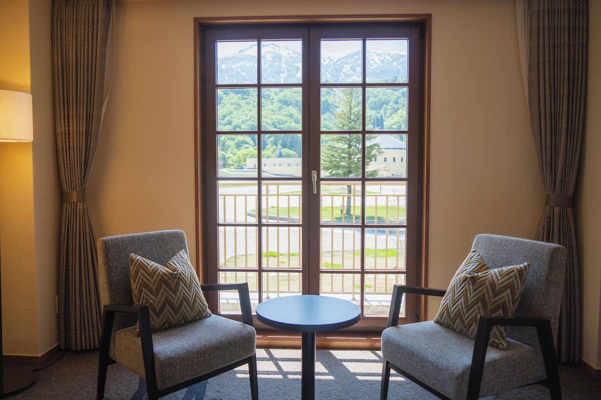 A room with two armchairs, a small round table, and a large window with a view of a building and greenery.