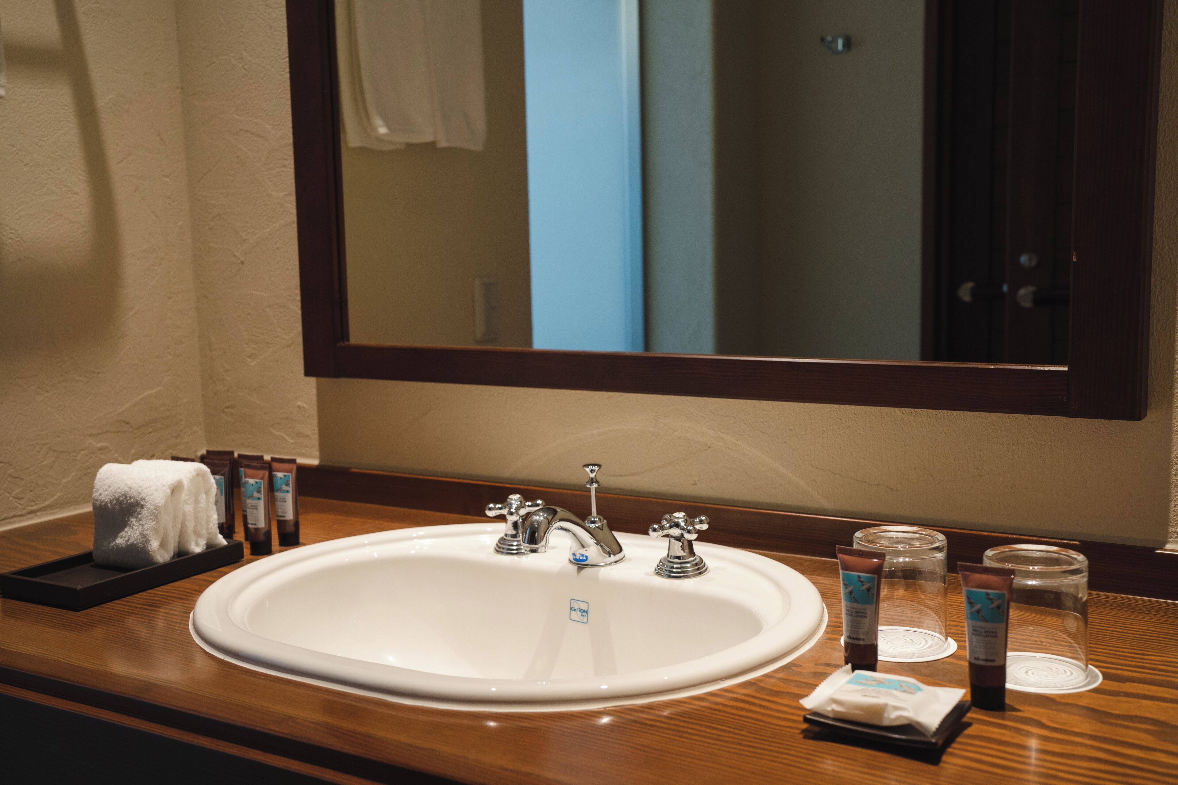 A hotel bathroom with a white sink, two faucets, a mirror, and toiletries on the counter.