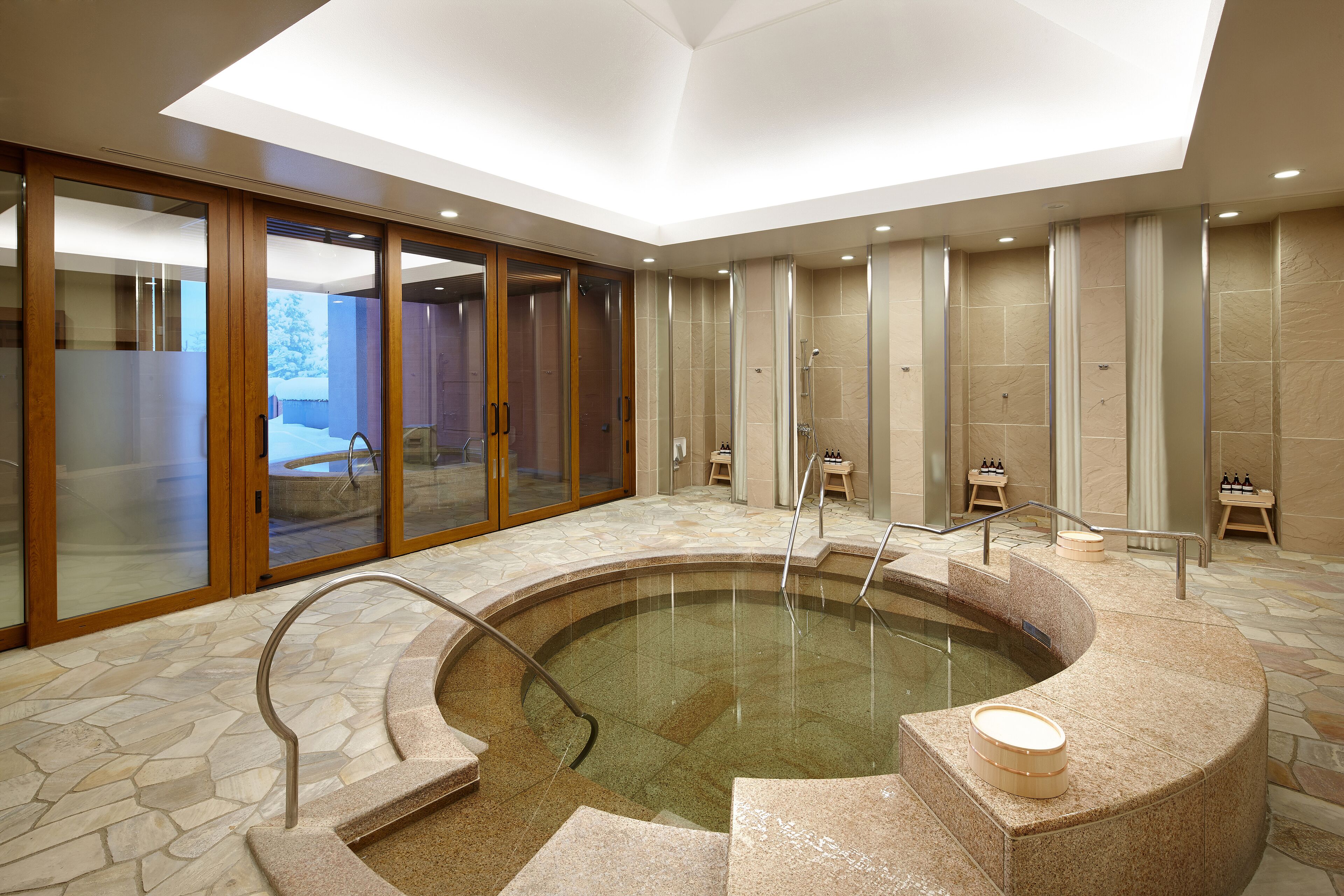 A modern indoor pool area with a stone floor, glass doors, and a seating area with a small table and chairs.