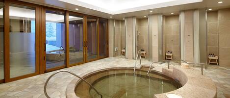 A modern indoor pool area with a stone floor, glass doors, and a seating area with a small table and chairs.