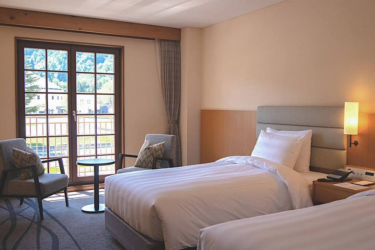 A hotel room with two beds, a chair, a small table, and a window with a view of greenery.