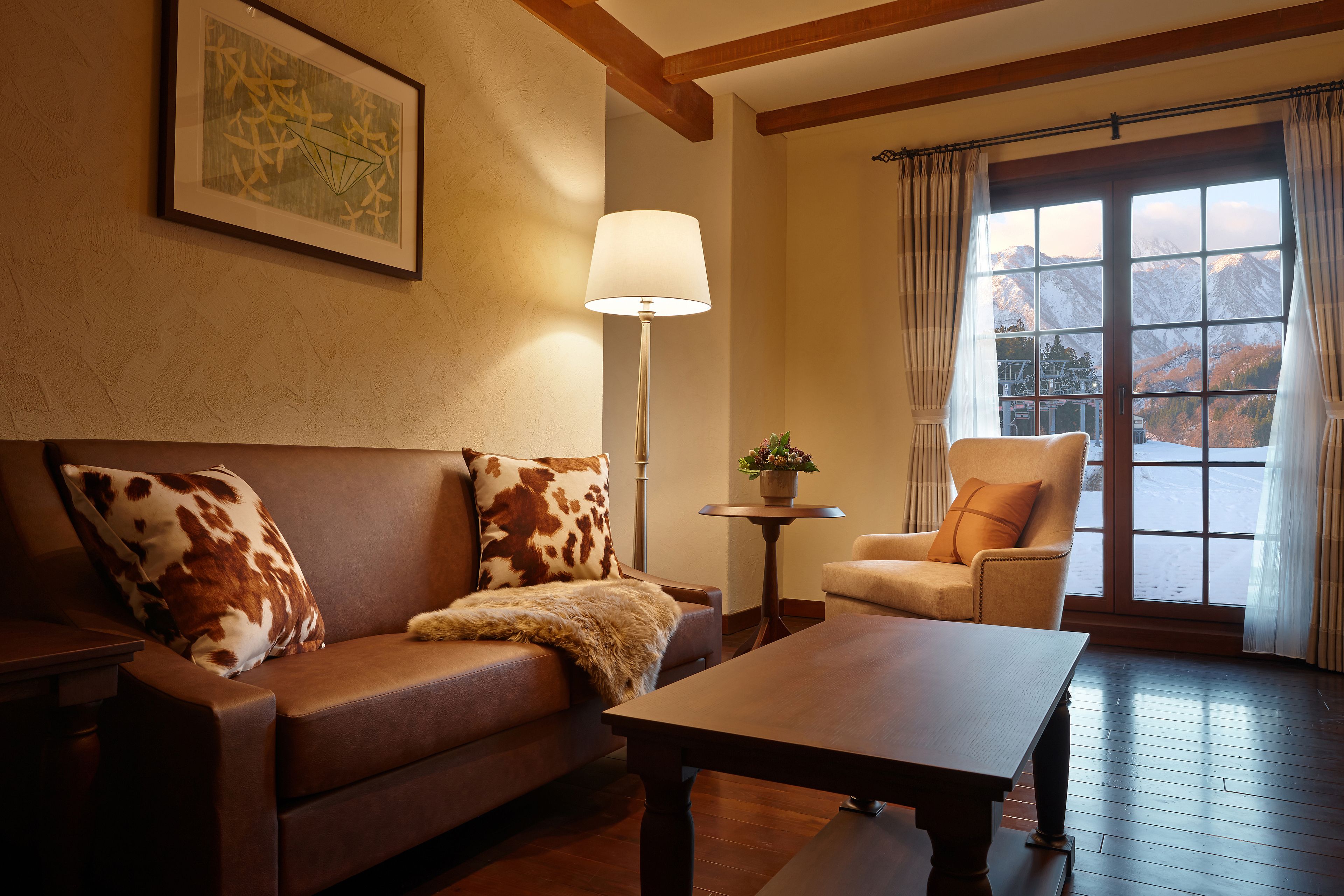 A cozy living room with a brown leather sofa, a wooden coffee table, and a view of mountains through a large window.