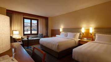 A hotel room with two beds, a desk with a chair, a small table, and a window with curtains.