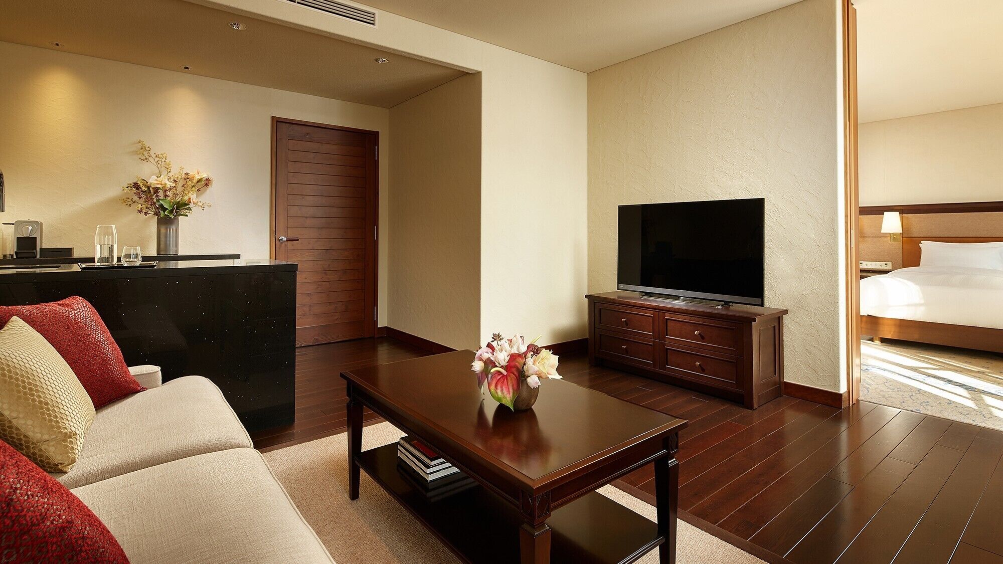 A hotel room with a sofa, a coffee table with a vase, a TV on a wooden stand, a bed, and a wooden door.