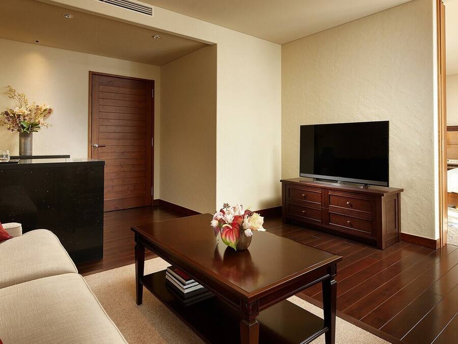 A hotel room with a sofa, a coffee table with a vase, a TV on a wooden stand, a bed, and a wooden door.