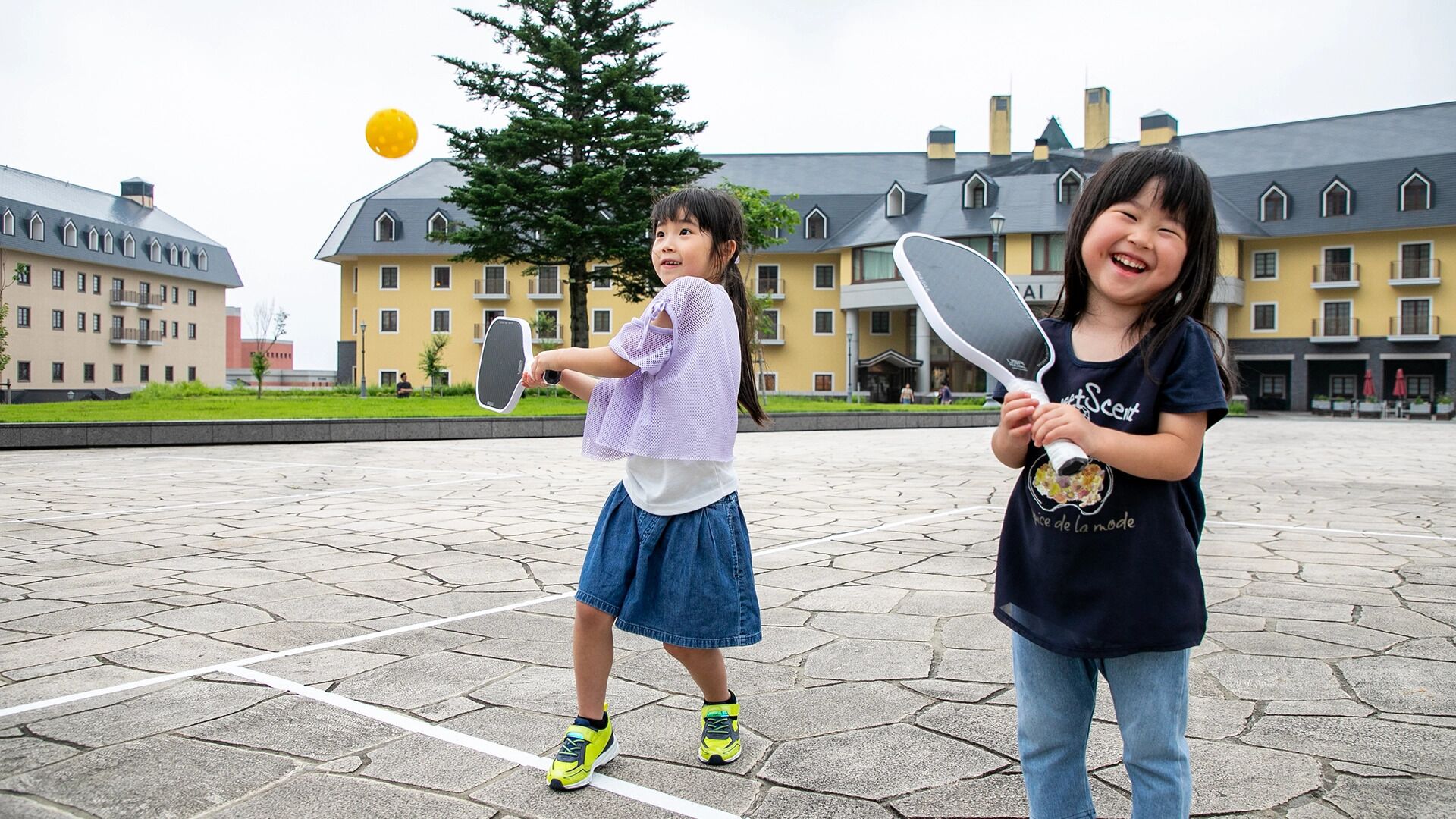 Two children playing tennis on a paved court with a building in the background.