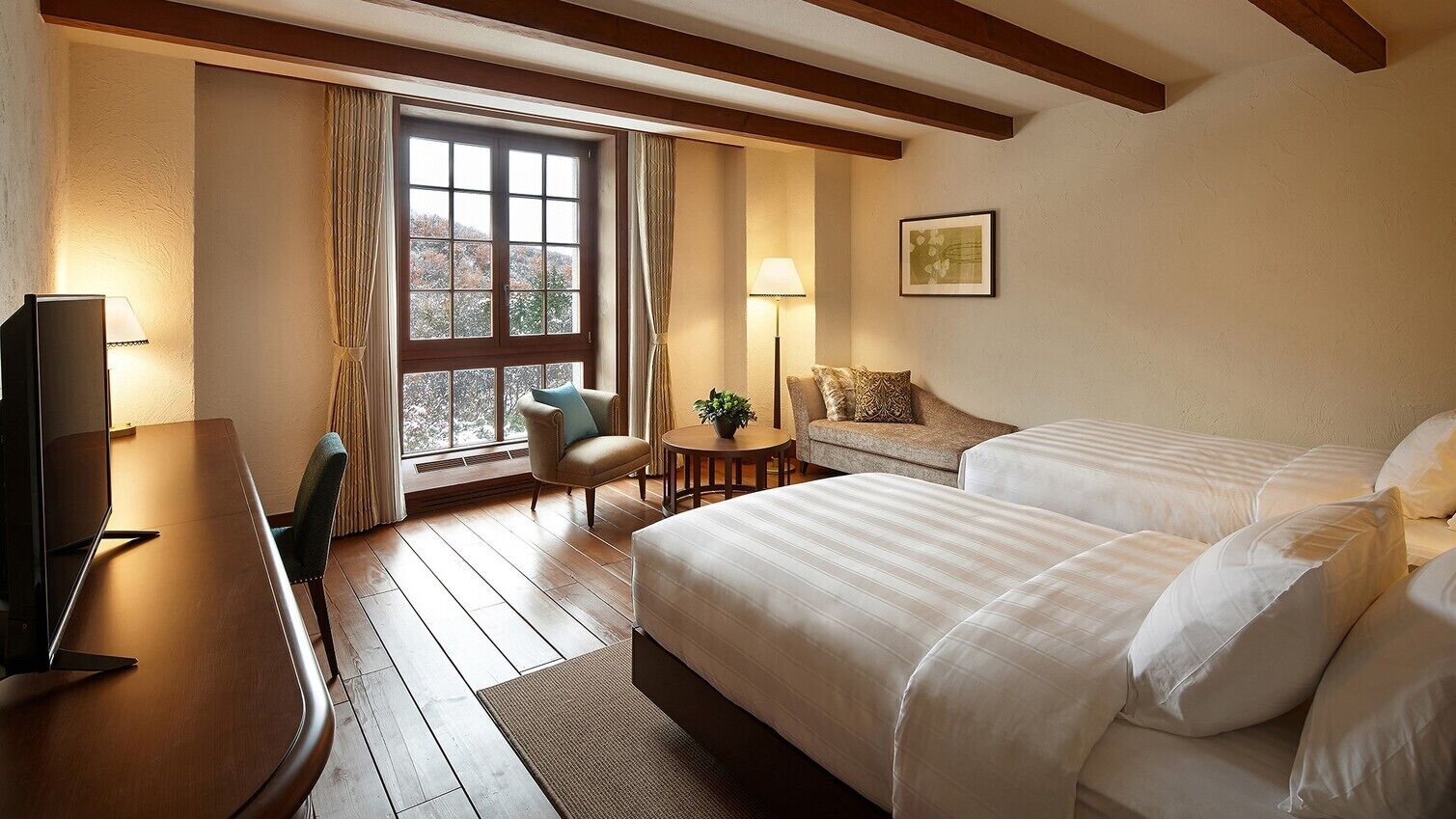 A hotel room with a wooden floor, a bed, a desk with a TV, a chair, a small table with a plant, and a window with curtains.