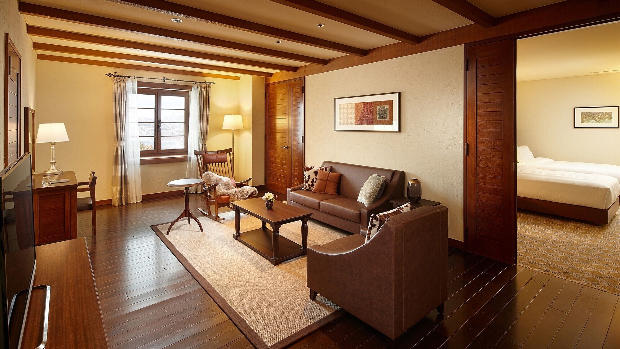 Habitación de hotel con piso de madera, un sofá, una mesa de centro, una silla, un escritorio, una lámpara, una cama y un cuadro en la pared.