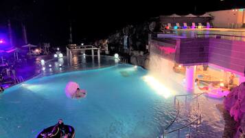 Relax by the illuminated pool, surrounded by lounge chairs and umbrellas, with a central water feature and a staircase leading to a higher level.