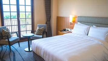 A hotel room with a large bed, two armchairs, a small table, and a nightstand with a lamp.