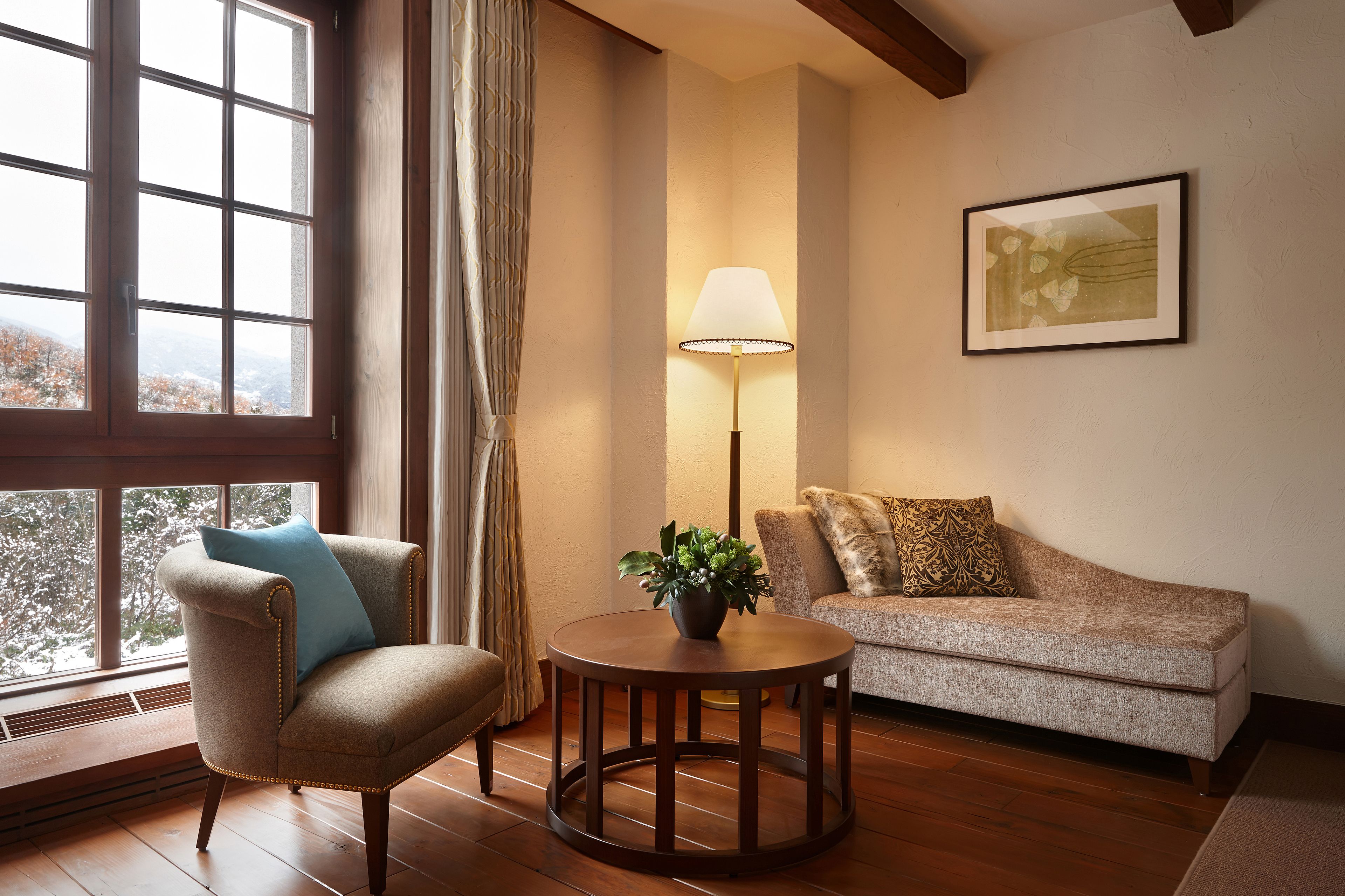 Una habitación acogedora con suelo de madera, una mesa redonda con lámpara, un sillón color beige y un sofá con cojines estampados.