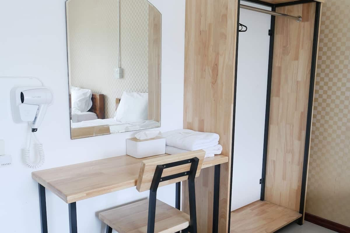 A compact hotel room with a wooden desk, a chair, a mirror, a small bedside table, and a foldable wooden wardrobe.