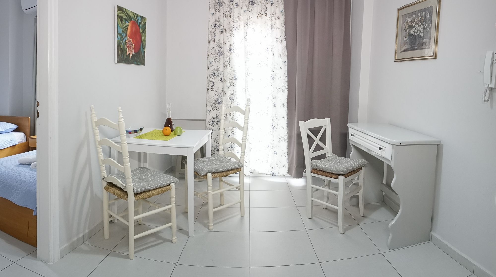 A small, clean room with a bed, a desk, two chairs, a table with fruits, and a window with curtains.