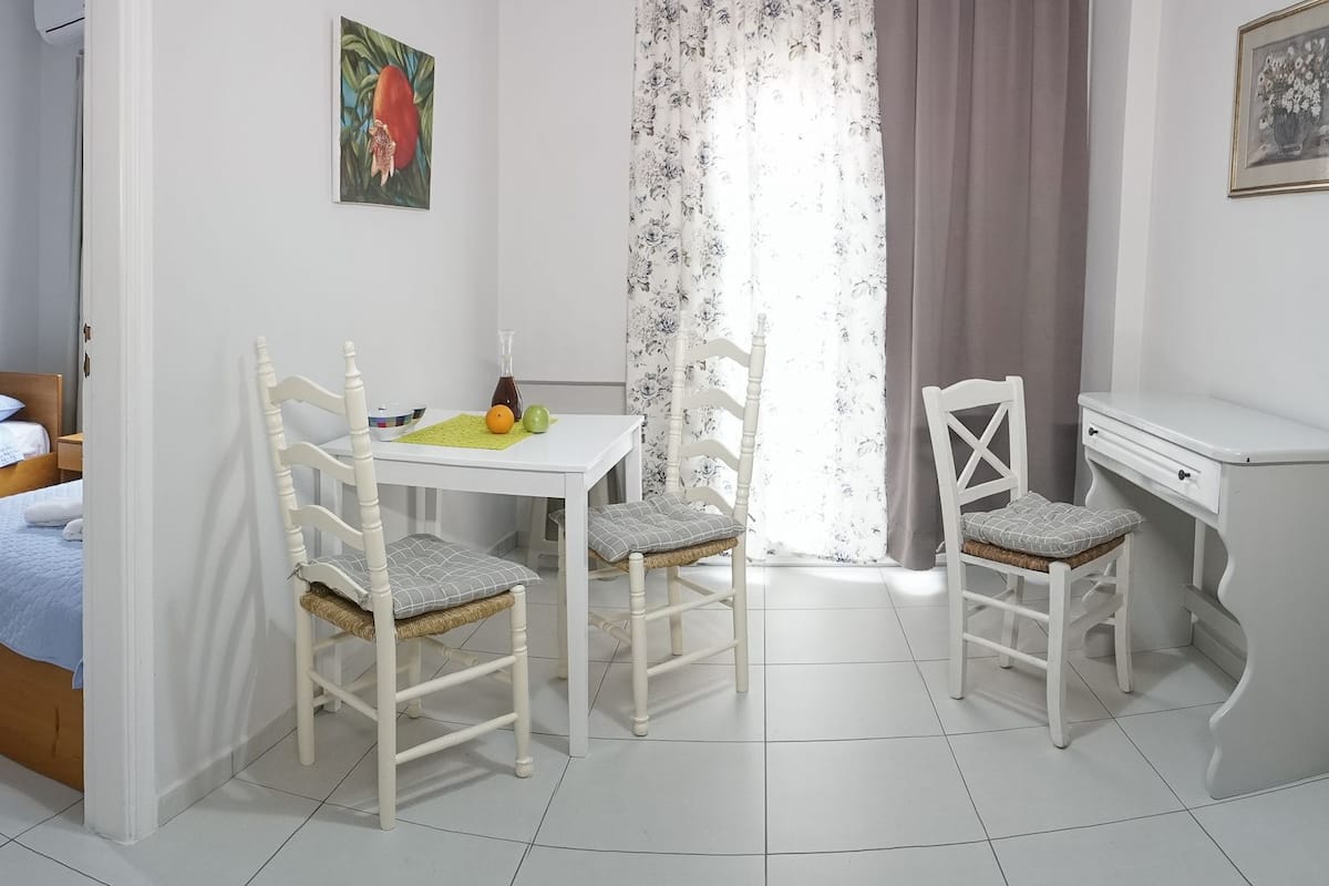 A small, clean room with a bed, a desk, two chairs, a table with fruits, and a window with curtains.