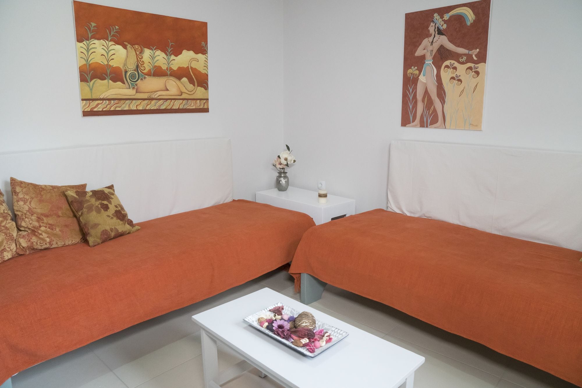 A room with two orange beds, a white coffee table with a floral arrangement, and two paintings on the walls.