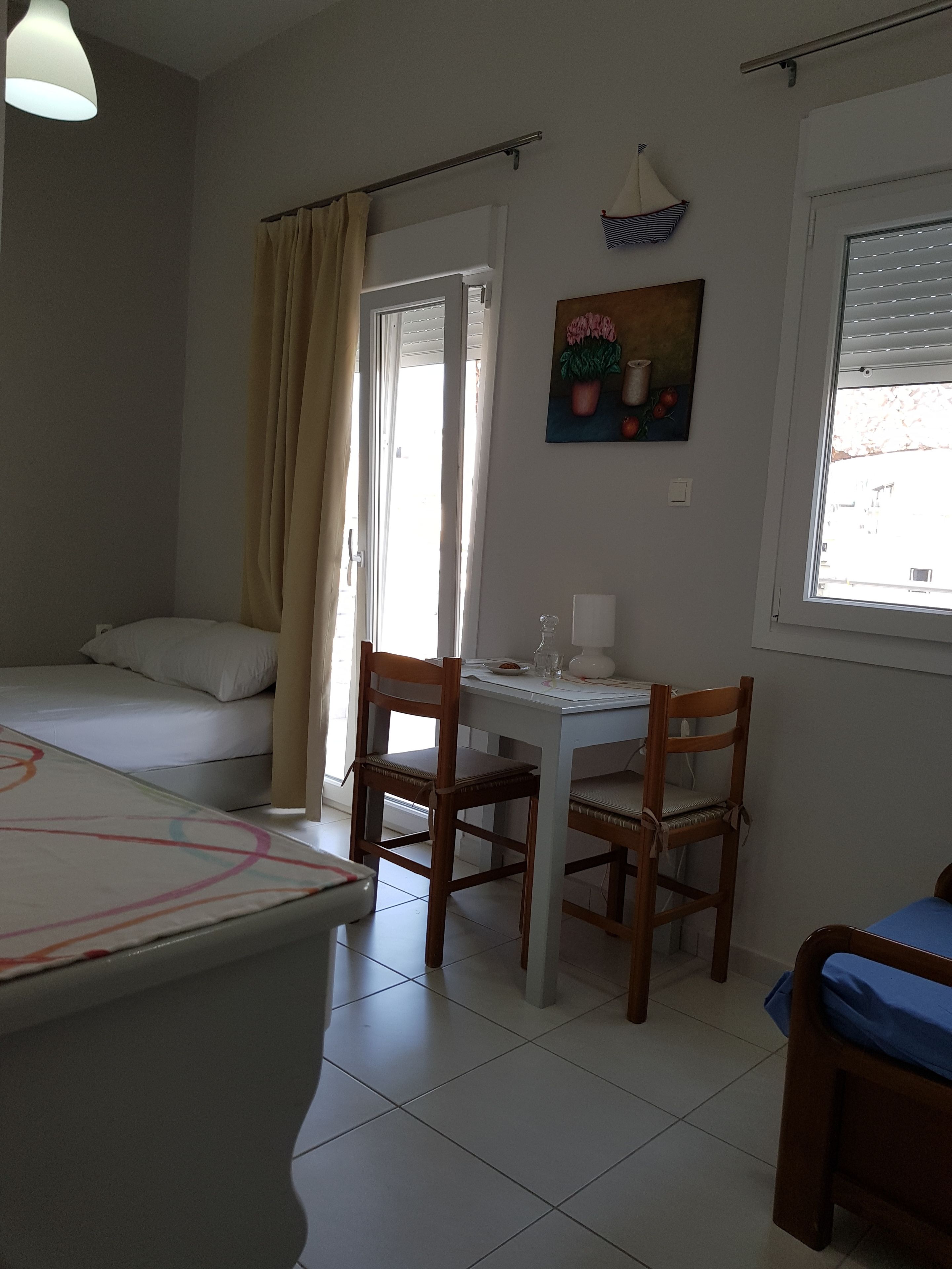 A room with a bed, a small table with chairs, a window with blinds, and a painting on the wall.