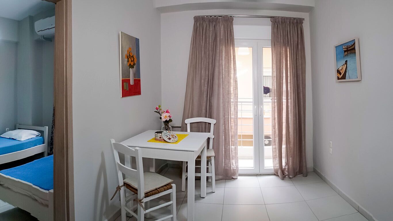 Una habitación pequeña con cama, una mesa y sillas, un ventanal con cortinas y obras de arte en las paredes.