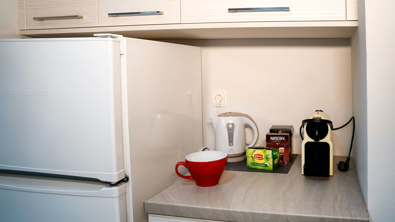 Una kitchenette compacta con un refrigerador blanco, una cafetera, un hervidor y una taza roja sobre la encimera.