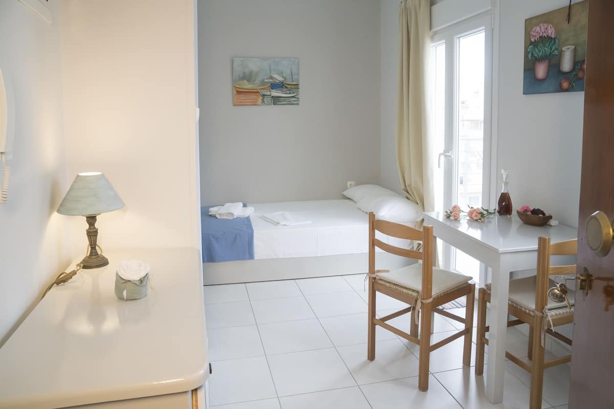 A small, clean room with a bed, a desk, and a chair. There is a painting on the wall, a lamp, and a window with curtains.