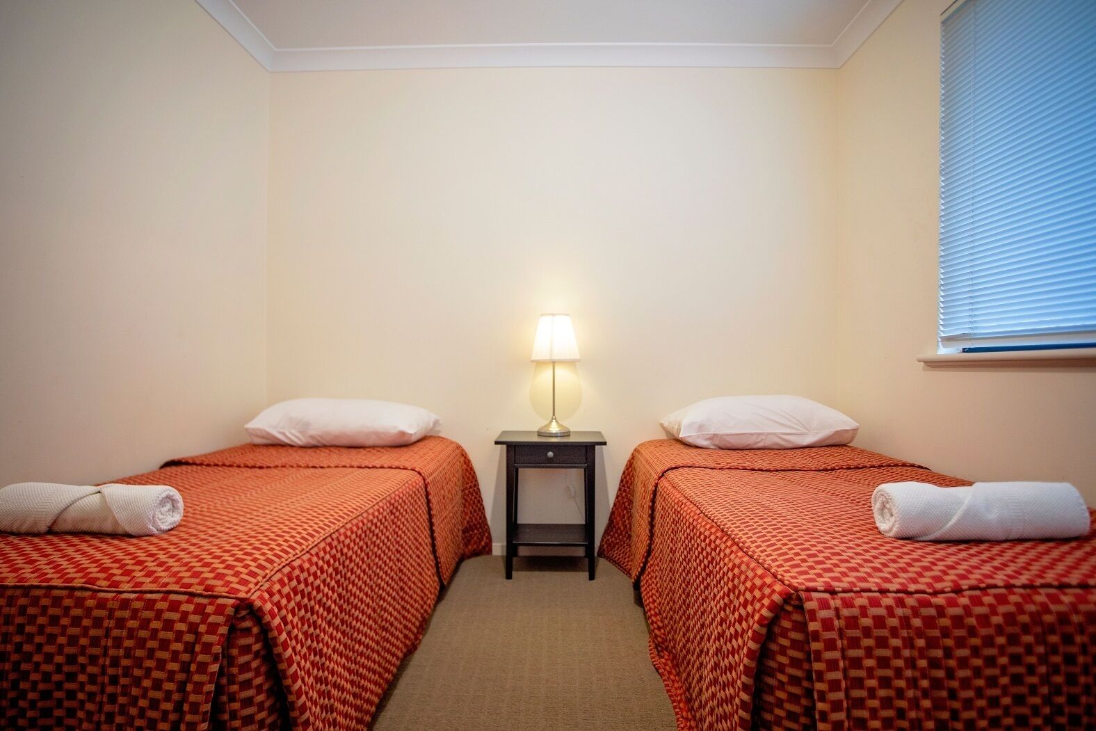 A hotel room with two single beds, each with a red checkered bedspread and white pillows. A small bedside table with a lamp is between the beds.