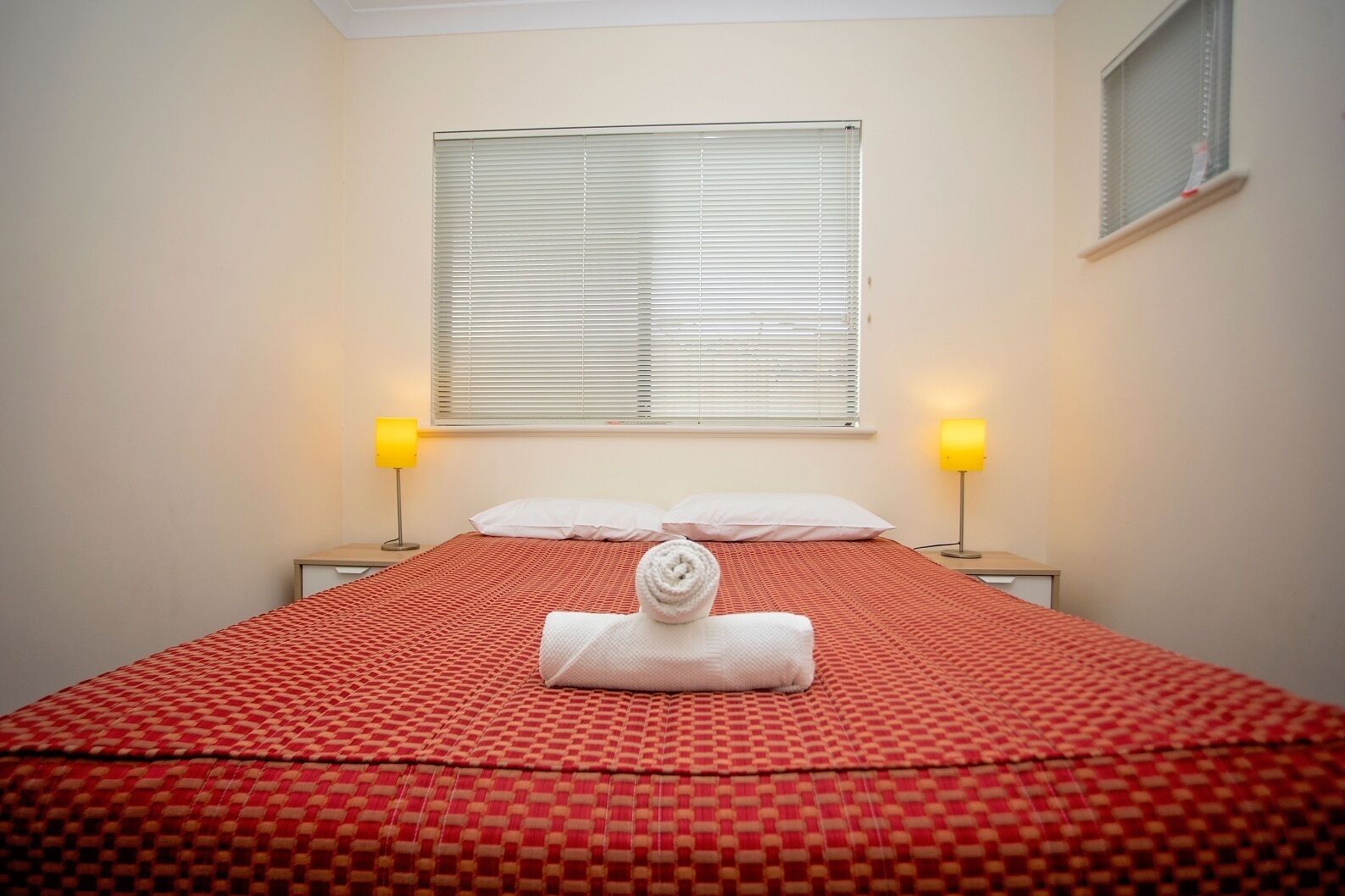 Habitación de hotel con cama, dos lámparas de noche, una ventana con persianas y una toalla sobre la cama.