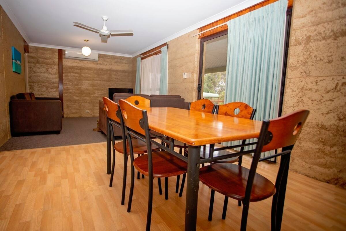 Un coin repas avec une table et des chaises en bois, un ventilateur de plafond et une vue sur l’extérieur à travers une fenêtre ornée de rideaux.