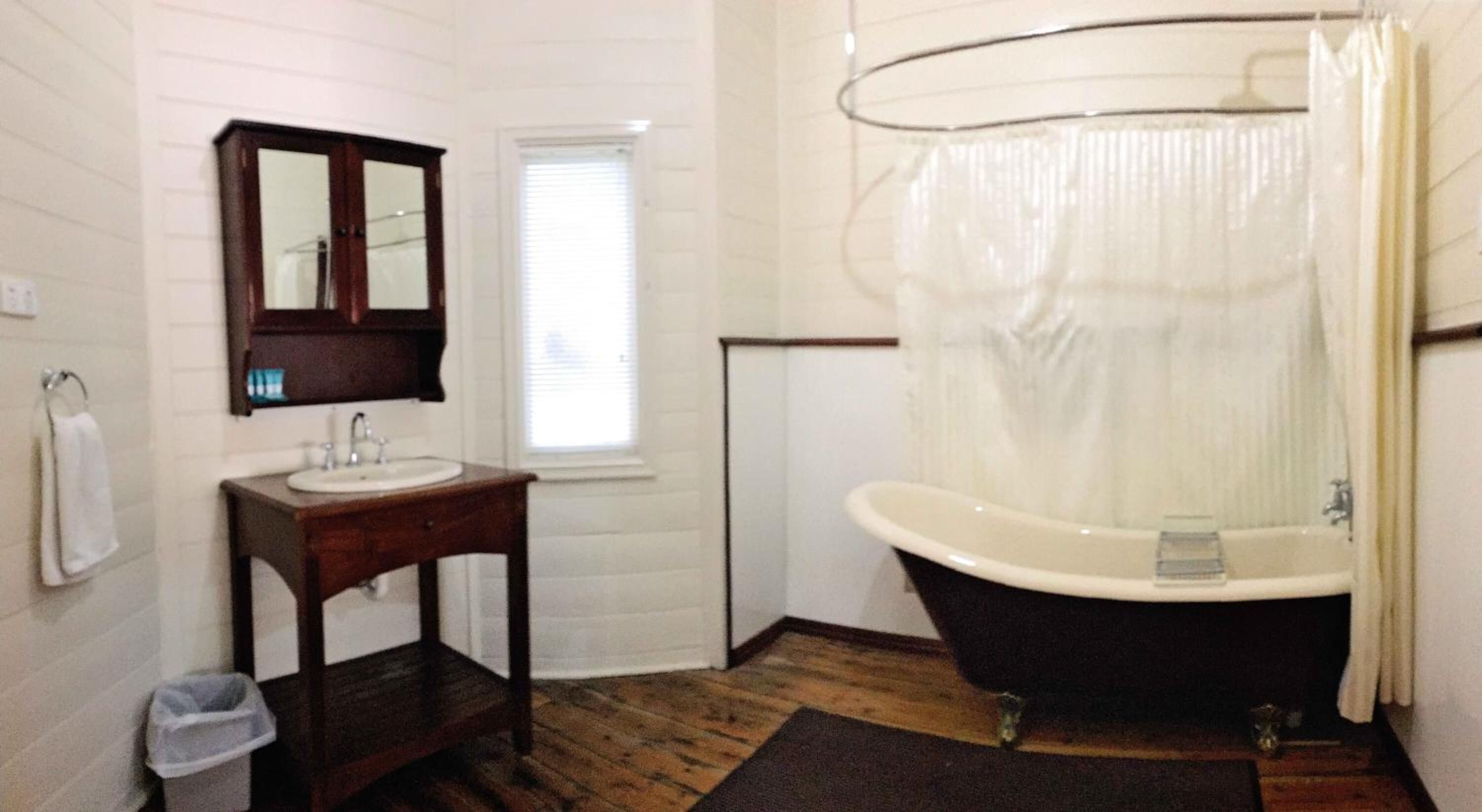 Un baño con tocador de madera, lavamanos blanco, espejo, bañera con cortina de ducha y ventana con persianas.