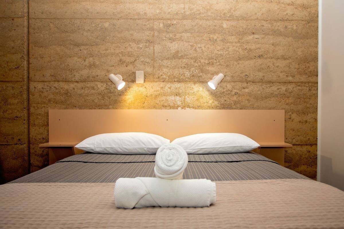 Un lit bien fait, avec du linge de lit blanc, une serviette enroulée et deux lampes fixées au mur.