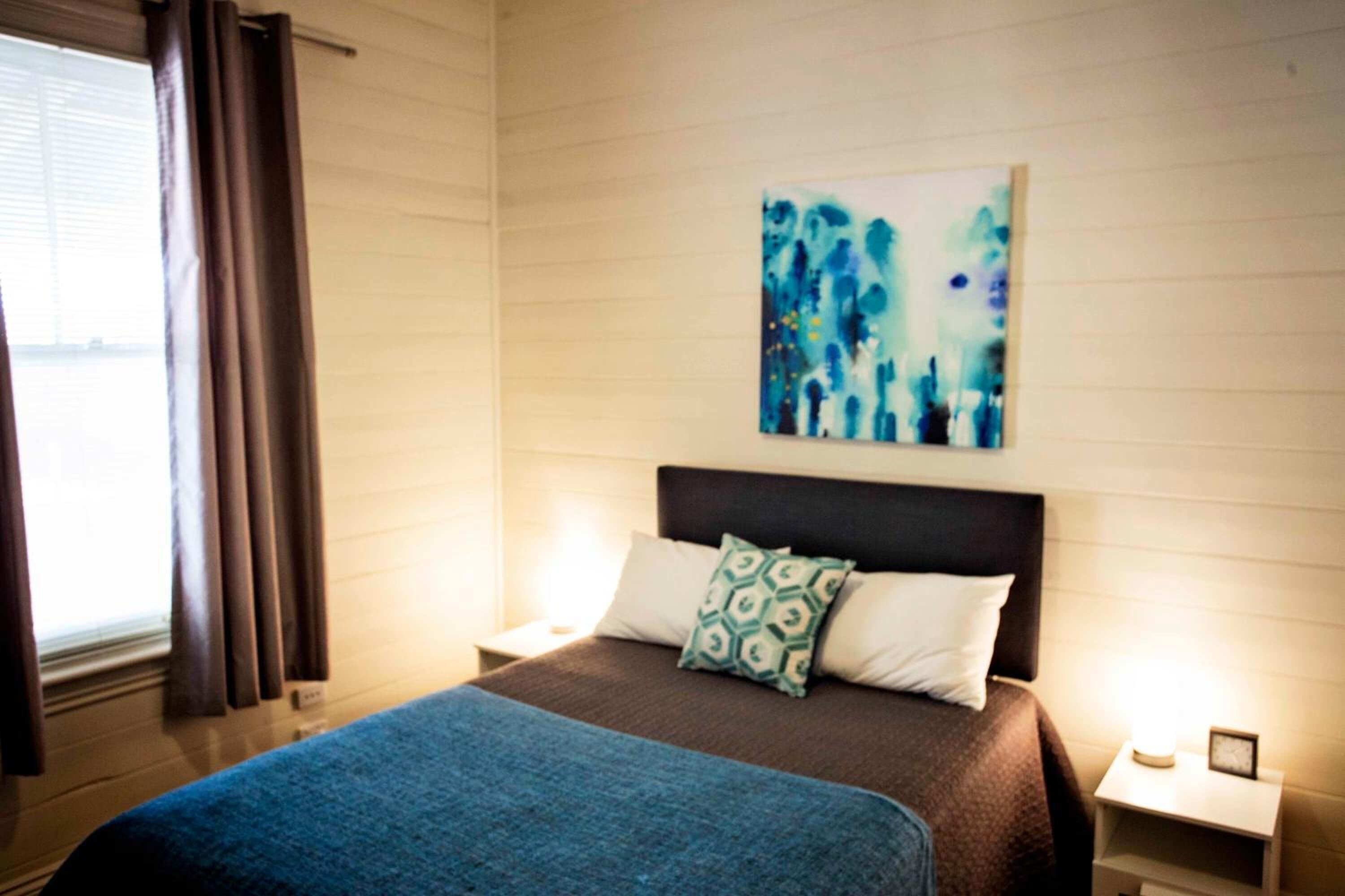 A hotel room with a bed, a nightstand, a wall-mounted painting, and a window with curtains.