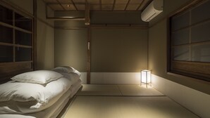 A traditional Japanese room with tatami flooring, a low bed with white bedding, a small bedside table with a lamp, and sliding doors.