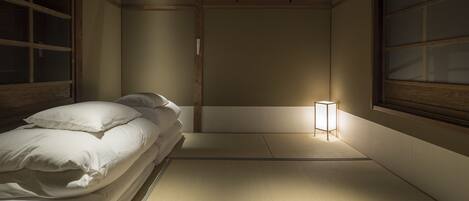 A traditional Japanese room with tatami flooring, a low bed with white bedding, a small bedside table with a lamp, and sliding doors.