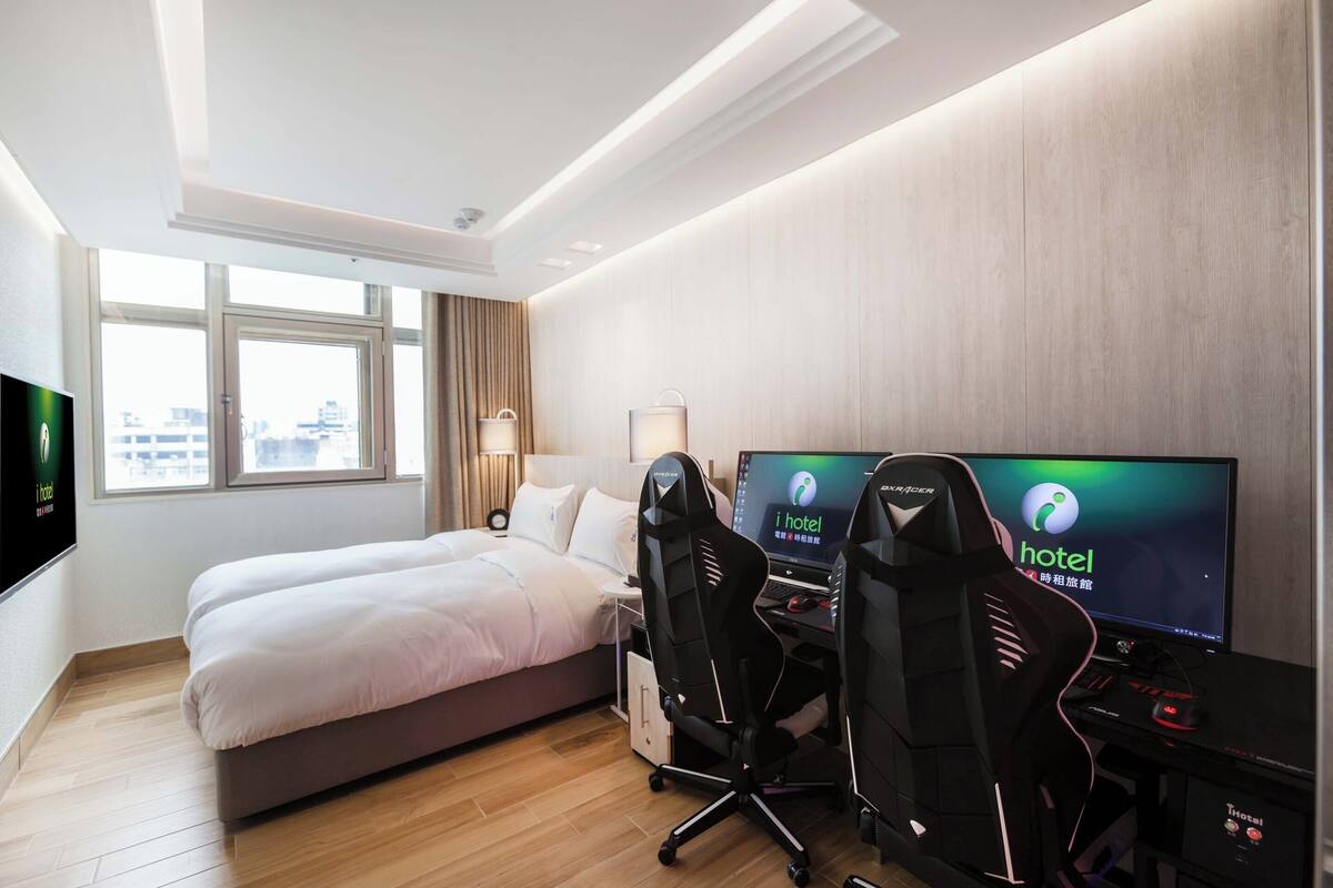 A hotel room with a bed, two gaming chairs, a desk with a computer, and a TV displaying 'i hotel'.