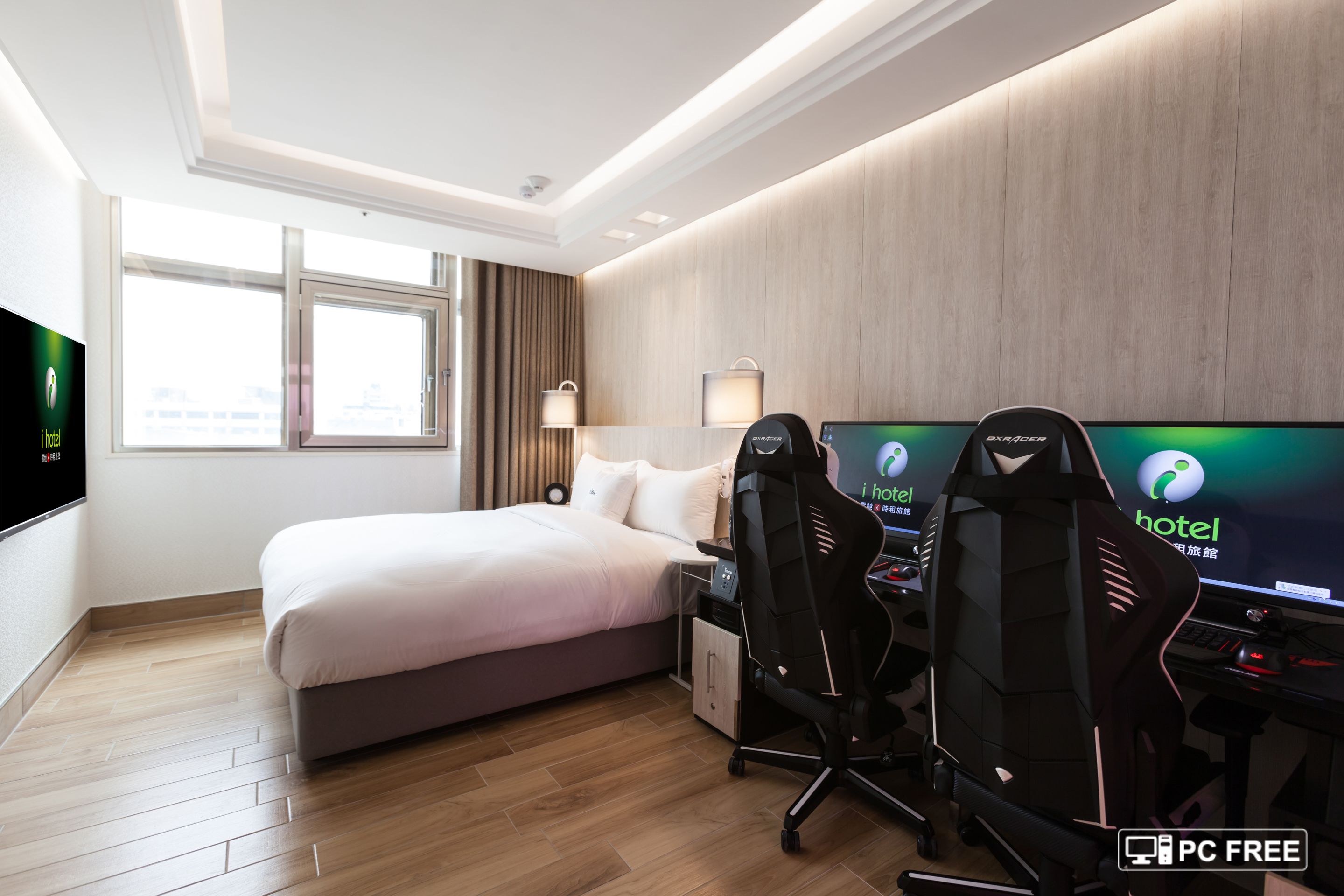 A hotel room with a bed, two gaming chairs, a desk with a computer, and a TV displaying the hotel's logo.