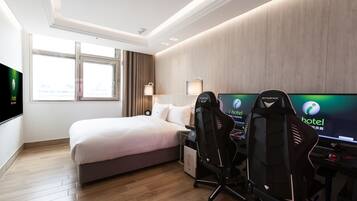 A hotel room with a bed, two gaming chairs, a desk with a computer, and a TV displaying the hotel's logo.