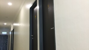 Un pasillo con tres puertas, cada una con un número. Las paredes son blancas y el suelo está cubierto de baldosas.