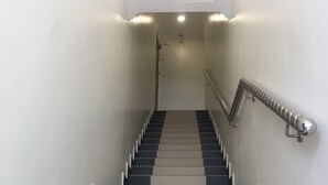 Una escalera con escalones alternados de color negro y beige, barandales metálicos y una pared blanca con una luz en el techo.