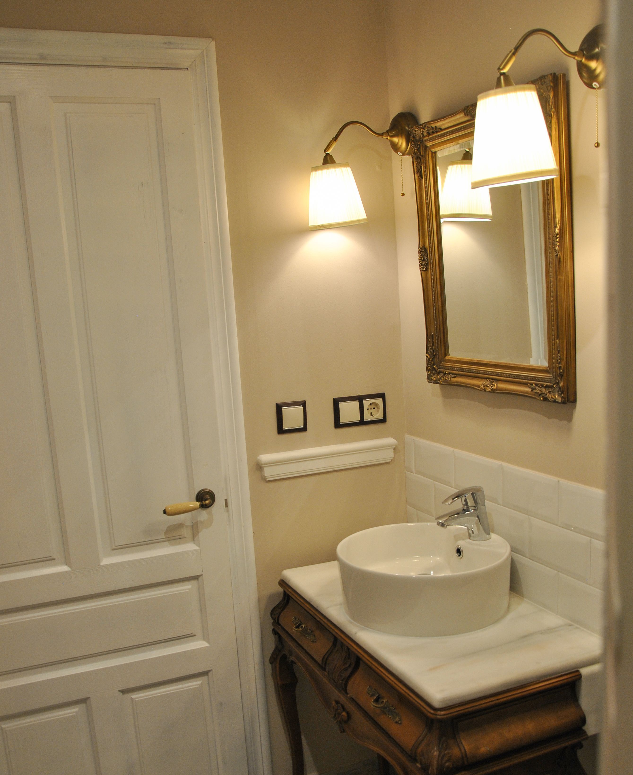 Un baño con una puerta blanca, un lavabo redondo con un solo grifo, un espejo con marco dorado y dos luces fijadas en la pared.