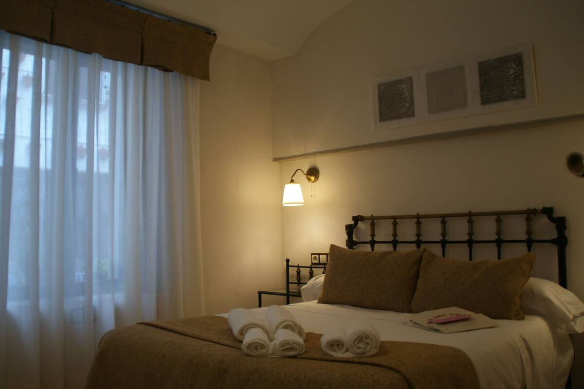 A hotel room with a bed, a nightstand, a wall-mounted lamp, and a window with curtains.