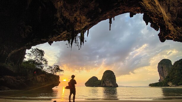 Eine Person steht an einem Strand mit einer Höhle im Hintergrund und blickt auf das Meer und ferne Inseln.