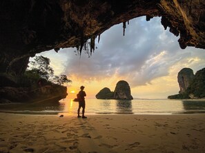 A person standing on a beach with a cave opening behind them, overlooking the sea and distant islands.